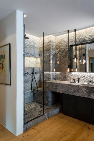 Das angrenzende Badezimmer ist durch das Spiel mit verschiedenen Naturmaterialien das Highlight der Wohnung. Als Raumteiler dient eine freistehende Badewanne, die mit schwarzem Naturstein einen Kontrast zum Holzboden bildet.