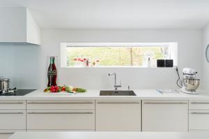 Die Fronten der Poggenpohl Küche ebenso wie die Arbeitsplatte aus Kunststoff sind einheitlich in blütenweiß gehalten. Die Edelstahlgriffe harmonieren mit dem warmen Holzboden.