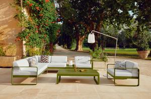 Modisches Fermob Outdoor-Möbelset, bestehend aus Bellevie Sofa, Balad Akkuleuchten, Alto Coffee Table, ergänzt durch Beistelltisch, niedrigen Tisch, Sessel und kreative Dekorationselemente wie Vase, Tablett und Bouquet Sauvage Kissen im Garten