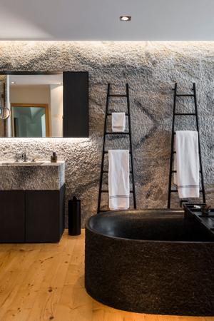 Das angrenzende Badezimmer ist durch das Spiel mit verschiedenen Naturmaterialien das Highlight der Wohnung. Als Raumteiler dient eine freistehende Badewanne, die mit schwarzem Naturstein einen Kontrast zum Holzboden bildet.